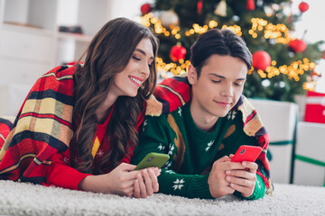 Fototapeta premium Portrait of two positive cheerful partners lay carpet floor under blanket hold use telephone christmastime indoors