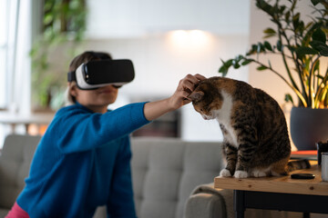 Positive woman in VR helmet petting cat sits on couch watching movie with 3D effects or virtual...