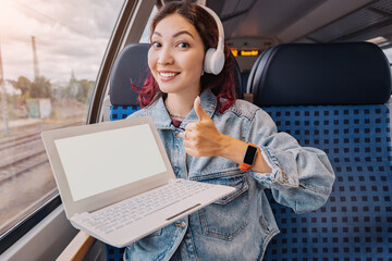 Happy young girl rides in a modern intercity train and shows white screen of her laptop. Work...
