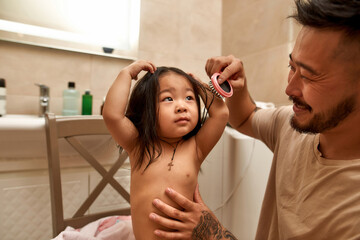Fototapeta premium Smiling asian father combing hair of daughter