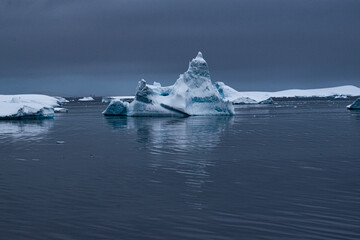 A snow-covered iceberg floating in the Antarctic Ocean, with a reflection in the water.  The dark grey sky is also reflected in the water. 