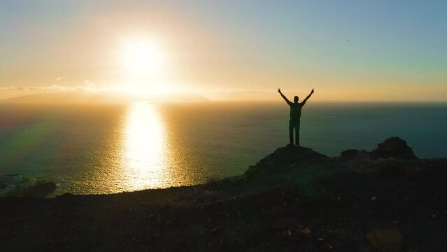 One travel man standing on top of crag at colorful sunset and raises hands up. Guy silhouette looking at orange sky with big sitting sun on ocean background. Adventure traveling hike concept.