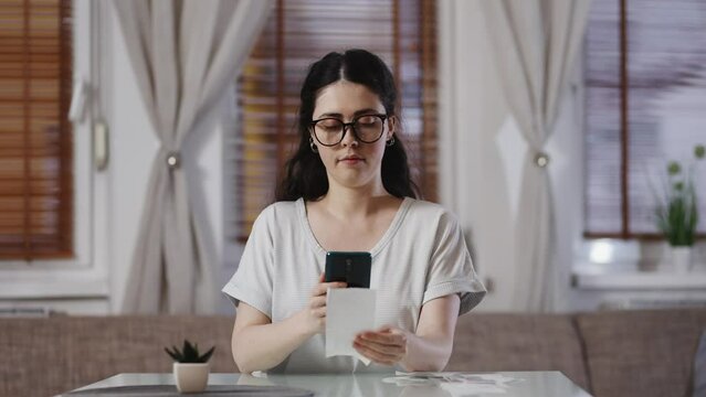 Young pretty Caucasian woman with glasses scans QR code on paper receipt from store with her smartphone. Concept of accounting and tax refund.
