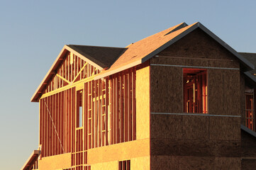 Two-story single-family home under construction with exposed wood framing and OSB sheathing, illuminated by morning sunlight, illustrating early-stage residential building progress