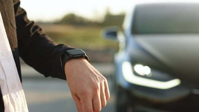 Unrecognizable elegant man using smart watch app for remote controlling of Tesla model x plaid electric car, driver distantly approaching car and opening door