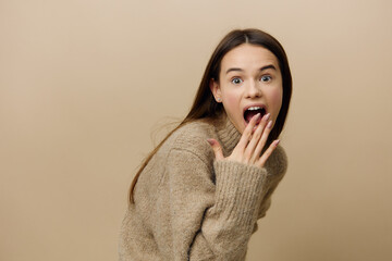a shocked, surprised woman looks into the camera with a very emotional face and covers her mouth with her hand, standing in a textured sweater on a beige background