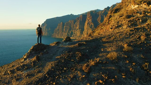Man backpacker stands on mountain top ocean coast at evening sky sunset background. Rear aerial view of tourist with backpack contempaltes rocky cliffs.