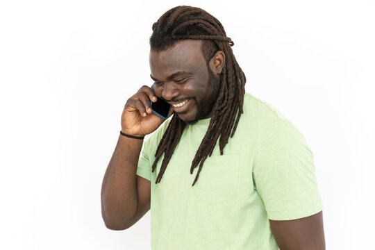 African American man with dreadlocks talking on phone, smiling. Black man standing against white background, looking down, having conversation with friend. Communication, happiness concept