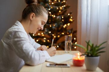 Indoor shot of young adult woman writing resolutions for New year in notebook at home, sitting by...