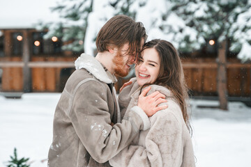 Fototapeta premium Young couple girl guy walking near barn with skates. Snowy cold winter in countryside. Skating, kissing, having fun, laughing in stylish clothes, fur coat,hat.Romantic love story,village date,weekend