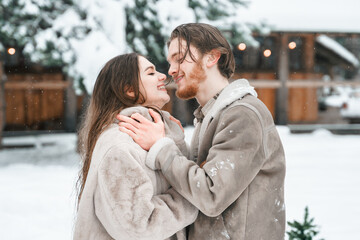 Young couple girl guy walking near barn with skates. Snowy cold winter in countryside. Skating, kissing, having fun, laughing in stylish clothes, fur coat,hat.Romantic love story,village date,weekend