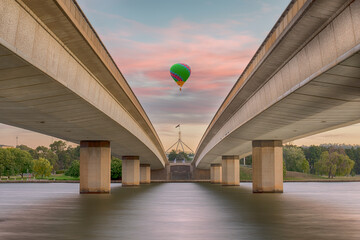 Unidentified hot air balloon passing over the commonwealth bridge, Canberra. with capital hill in the background during sunrise. - obrazy, fototapety, plakaty