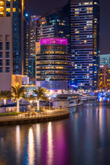 Night view to Dubai Marina skyline, reveals Pier 7 and boats. Luxury destination for tourists and residents. Amazing colors reflect on the water.