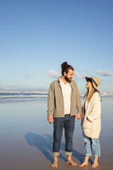 Thrilled couple walking on beach. Bearded man and woman in casual clothes looking at each other, holding hands. Love, vacation, affection concept