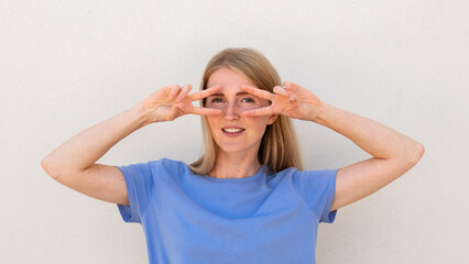 Cheerful young woman showing V-signs near her eyes. Caucasian female model with blonde hair and blue eyes in blue T-shirt smiling and holding victory signs near eyes. Posing, entertainment concept