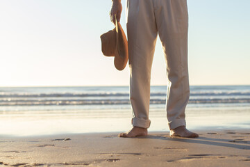 Closeup of barefoot Caucasian mans legs standing at the beach, holding straw hat in his hand. Lower male body part in beige trousers on wet sand against sea background. Holiday, vacation concept