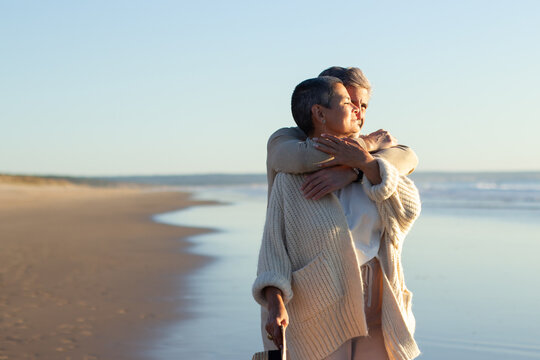 Romanic senior couple spending time at seashore at sunset, enjoying fantastic sea view. Grey-haired man hugging his wife while standing behind. Romance, retirement, holiday concept