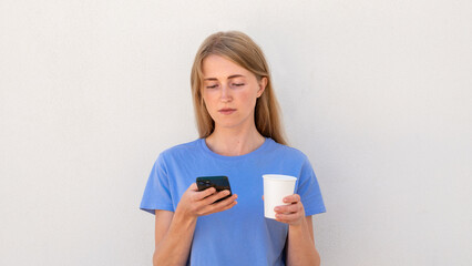 Thoughtful young woman using smartphone in her hand. Caucasian female model with blonde hair and blue eyes in blue T-shirt reading messages or news on smartphone. Modern technology concept