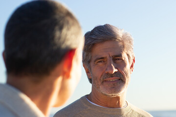 Over-the-shoulder shot of grey-haired senior man with beard talking to short-haired person outside. Portrait of happy calm man at seaside. Leisure, lifestyle concept
