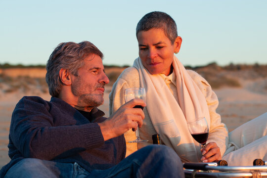 Beautiful senior couple drinking red wine at seashore at sunset while having picnic and enjoying warm evening at seaside. Short-haired lady and bearded man relaxing outdoors. Romance, leisure concept