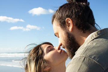 Fototapeta premium Passionate young couple spending time at seashore on sunny day. Closeup of couples faces close to kissing, touching with their noses against sea background. Romance, love concept