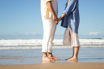Caucasian couple standing barefoot at seashore on wet sand holding hands. Slim woman in long blue shirt and man in white trousers enjoying vacation together. Sea background. Love, holiday concept