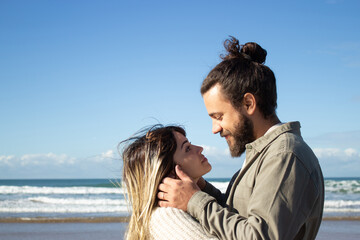 Happy romantic couple spending time at seashore, showing affection and hugging. Bearded guy with messy hairstyle gently holding girls face and smiling while wind playing with her hair. Romance concept