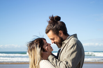Happy couple spending time at seashore, showing affection and embracing. Loving guy with beard tenderly holding girls face, while beautiful woman with closed eyes waiting for kiss. Romance concept