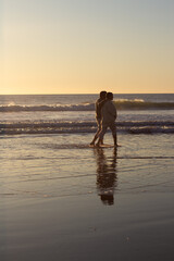 Senior couple strolling along seashore at sunset with their bare feet in water while waves rushing in background. Cinematic long shot. Romance, lifestyle, relationship concept