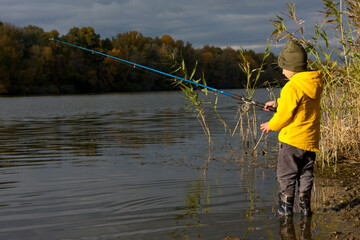 Young fisherman. boy in yellow jacket and rubber boots is fishing while standing in water on river bank. Leisure outdoors. gold autumn