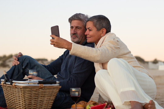 Lovely senior couple enjoying picnic at seashore in evening, drinking wine and looking through photos on mobile phone. Grey-haired man smiling while lady holding cellphone. Technology, romance concept