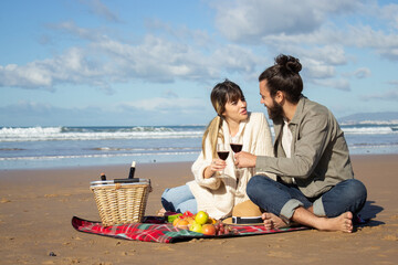 Young Caucasian couple drinking red wine at seashore enjoying romantic picnic. Pretty blonde lady and handsome guy clinking glasses while waves rushing to shore in background. Romance, leisure concept