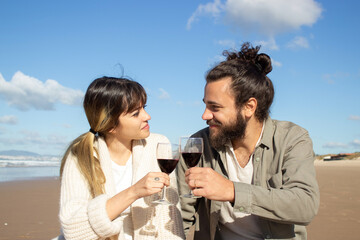 Romantic Caucasian couple drinking red wine at seashore enjoying picnic on sunny day. Pretty blonde lady and handsome guy clinking glasses and looking at each other. Romance, leisure concept