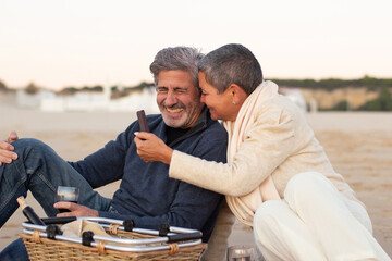 Romantic senior couple enjoying picnic at seashore, drinking wine and looking through photos on phone. Grey-haired man laughing while lady holding cellphone and smiling. Leisure, romance concept