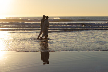 Senior couple walking along seashore at sunset with their bare feet in water while waves rushing in background. Cinematic long shot. Romance, lifestyle, relationship concept
