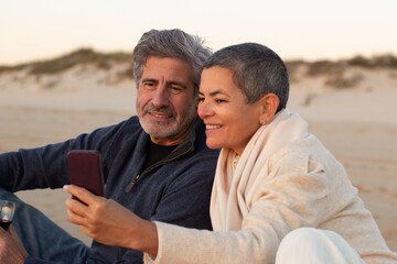 Senior couple spending time at the beach at sunset. Grey-haired man and smiling lady with short hair sitting on blanket and looking through photos on cellphone. Leisure, mobile communication concept