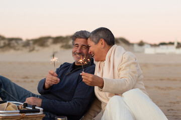 Senior couple enjoying picnic at seashore, holding sparklers and laughing. Lady with short hair and...