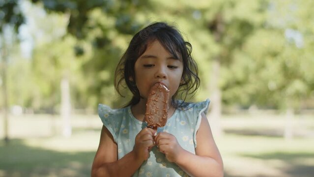 Portrait of cute little Asian girl eating choc ice outdoors. Toddler girl wearing blue dress eating ice cream. Childhood and sweet food concept