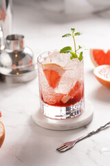 Preparation of a grapefruit alcohol free cocktail - A tumbler glass with ice cubes on marble surface with fresh mint surrounded by steel cocktail shaker and bar equipment