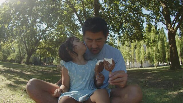 Portrait of happy Asian father and his daughter eating choc ice. Young man sitting with toddler girl on grass in park, child kissing her dad. Fatherhood and childhood concept