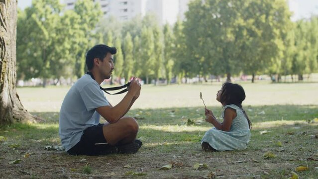 Happy Asian man taking photo of little daughter with camera. Toddler girl wearing dress sitting on ground holding twig. Fatherhood and childhood concept