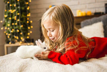 A little girl with a cute white rabbit is lying on the bed. Rabbit symbol of 2023