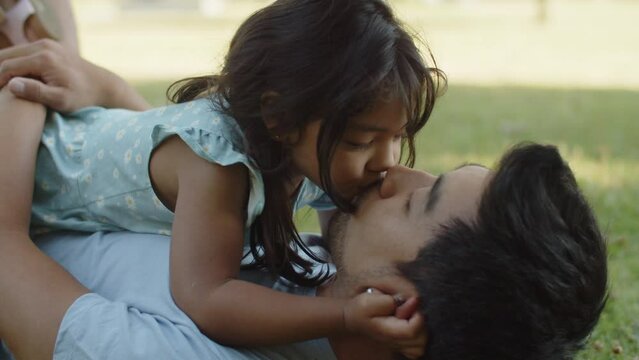 Little girl lying with father on ground and kissing him on lips. Asian young man resting on grass with little daughter in park. Fatherhood and childhood concept