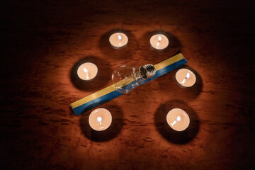 Power outage concept. An electric light bulb with a yellow-blue ribbon (the national symbol of Ukraine) in a circle of burning candles