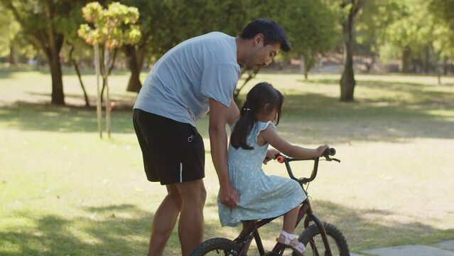 Happy Asian man teaching his little daughter to ride bike in park. Young father walking with toddler girl in summer. Fatherhood and childhood concept