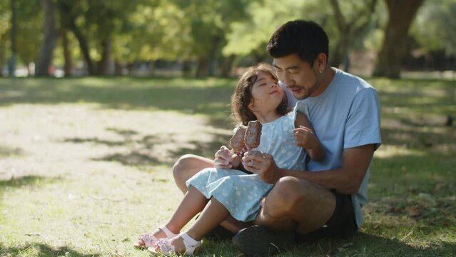 Cute little daughter sitting with father and eating choc ice in park. Young Asian man sitting on grass with child, girl kissing her dad. Fatherhood and childhood concept
