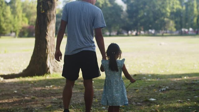 Rear view of man walking with toddler daughter in park. Cute Asian girl wearing blue dress. Fatherhood and childhood concept