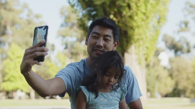 Happy Asian young father taking selfie with daughter in park. Smiling man sitting with child and taking photo with smartphone in park. Fatherhood and childhood concept