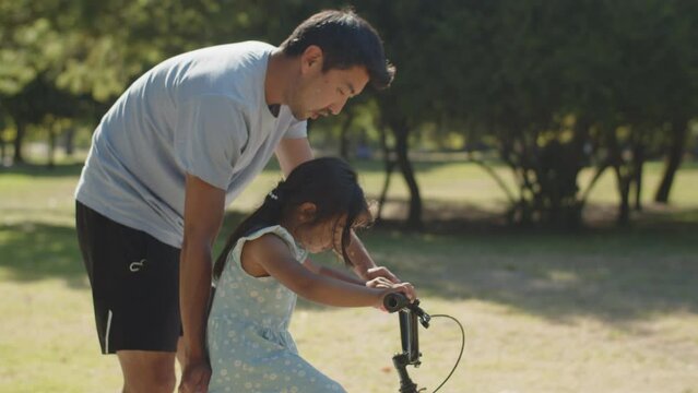 Happy young father teaching his daughter to ride bike in park. Toddler girl learning cycling with her dad in summer. Fatherhood and childhood concept