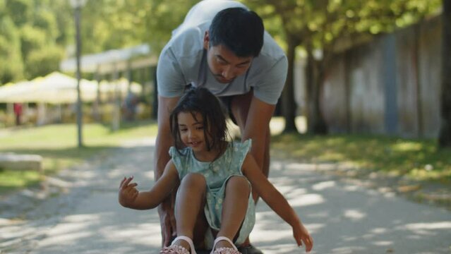 Young father giving a ride on skateboard to his little daughter in park. Happy toddler girl wearing dress sitting on board and smiling Fatherhood and childhood concept